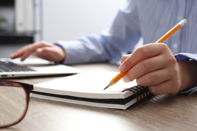 Woman with notebook working on laptop at wooden table in office, closeup