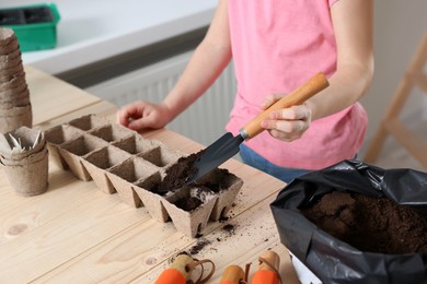 Little girl adding soil into peat pots at wooden table indoors, closeup. Growing vegetable seeds