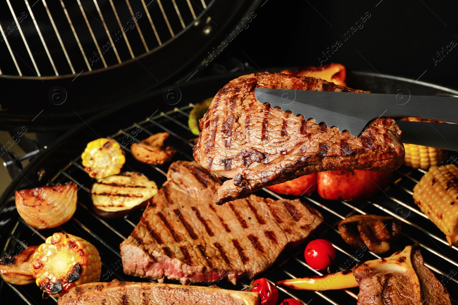 Photo of Tongs with fresh juicy meat steak over barbecue grill