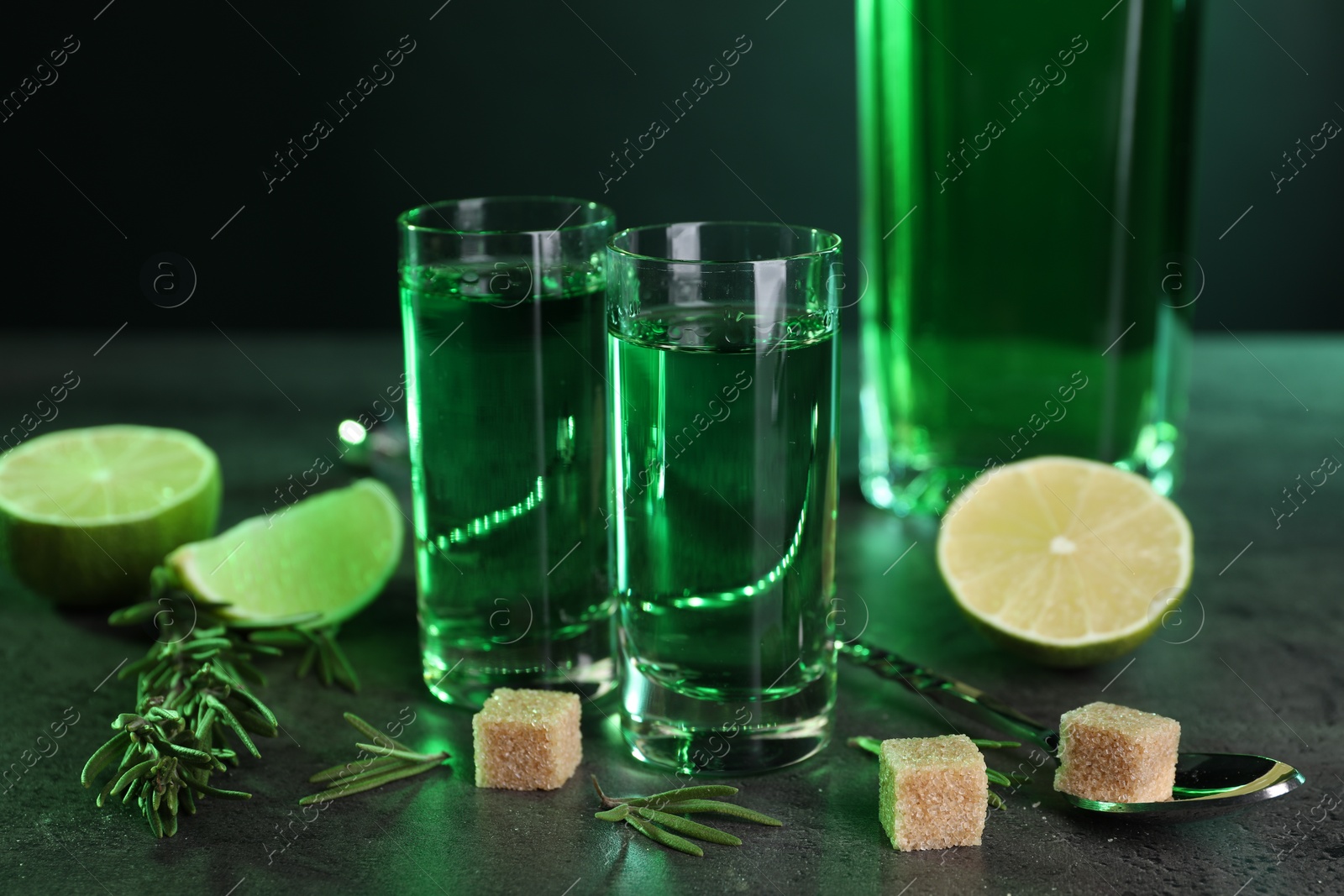 Photo of Absinthe in shot glasses, spoon, brown sugar, lime and rosemary on gray textured table, closeup. Alcoholic drink