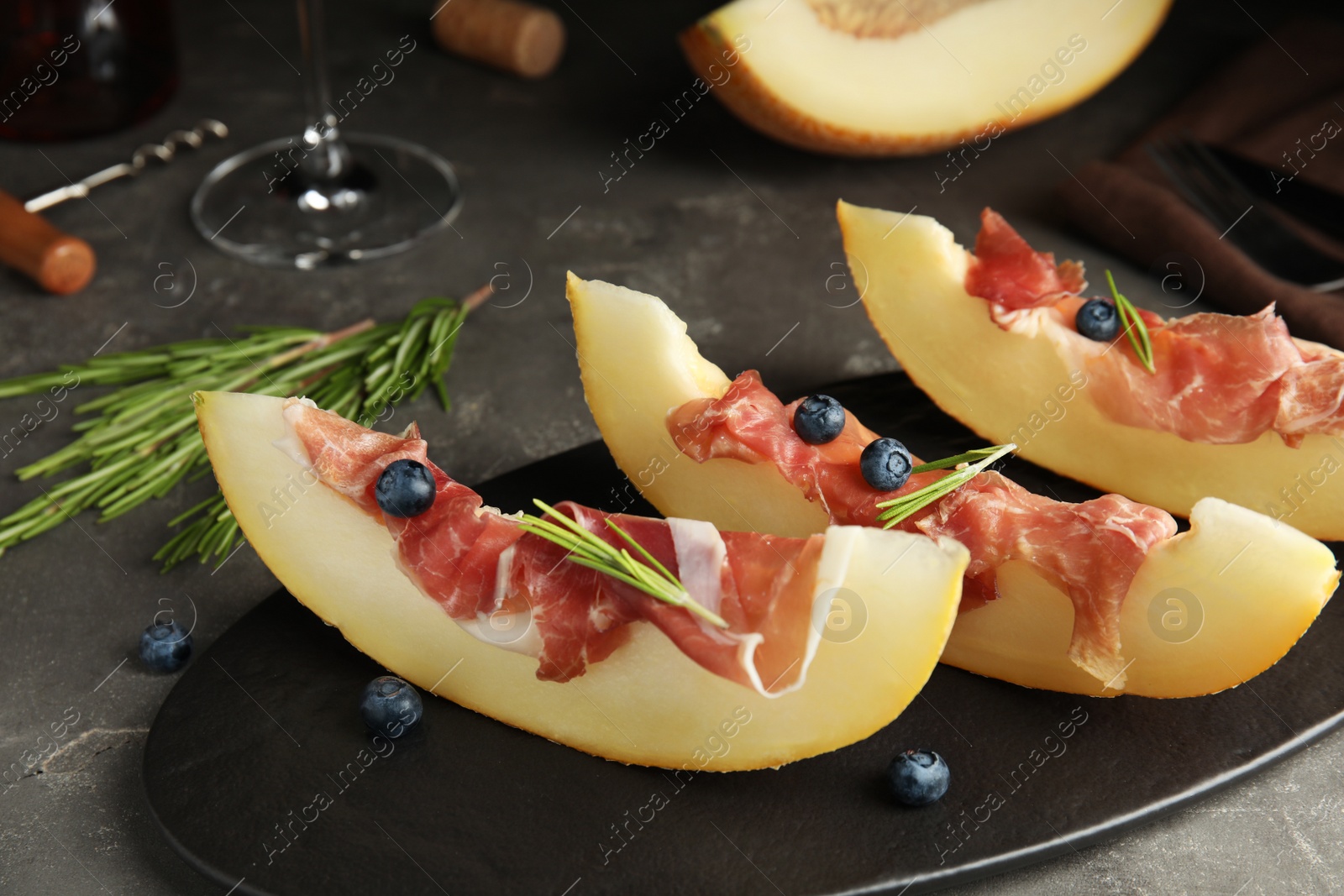 Photo of Melon with prosciutto, blueberries and rosemary on grey table