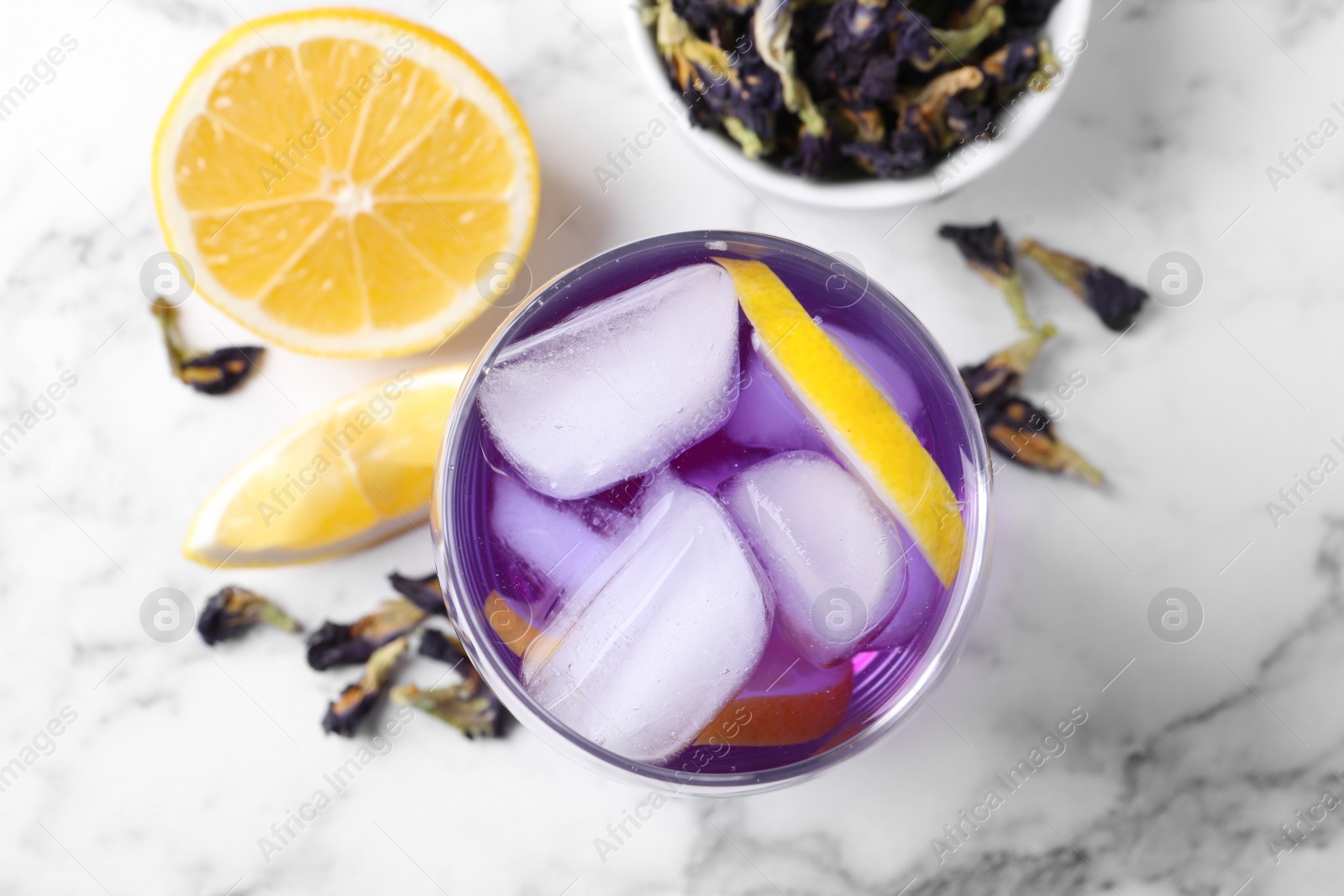 Photo of Glass of organic blue Anchan with lemon and ice on white marble table, flat lay. Herbal tea