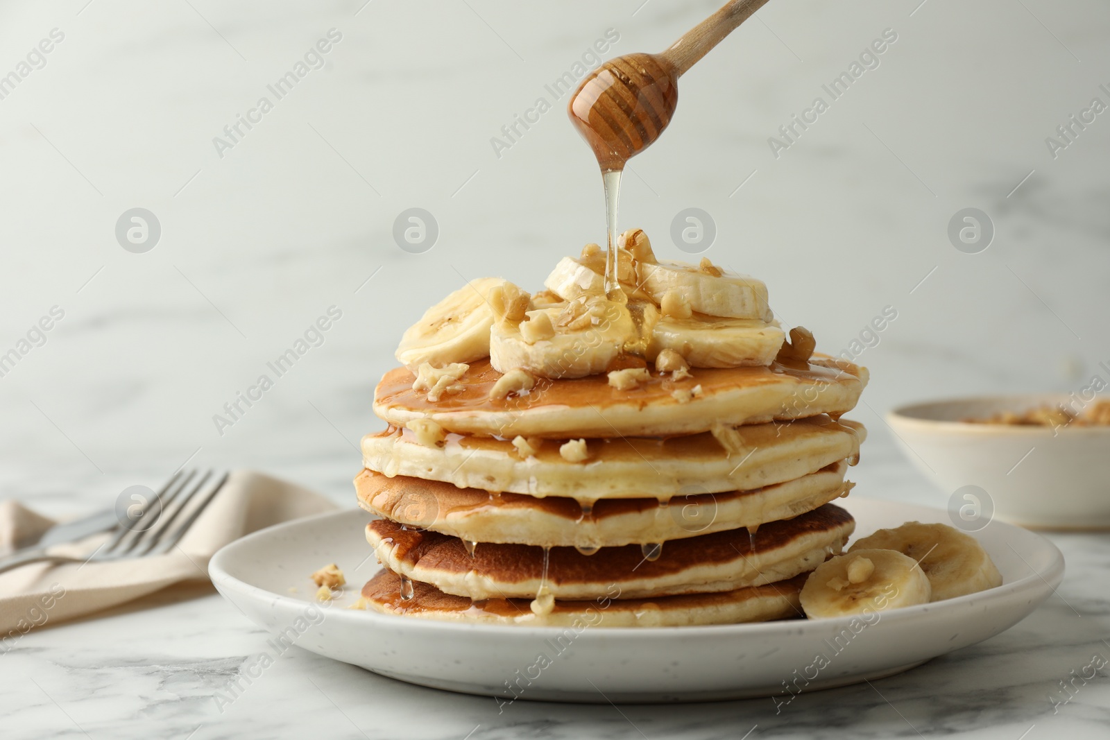 Photo of Pouring honey from dipper onto delicious pancakes with bananas and walnuts at white marble table, closeup