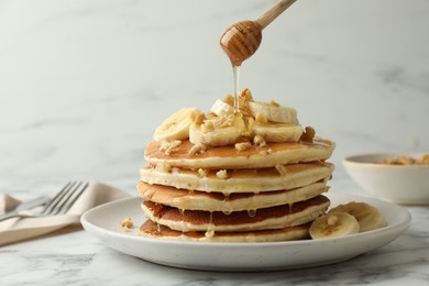 Photo of Pouring honey from dipper onto delicious pancakes with bananas and walnuts at white marble table, closeup