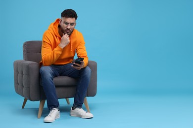 Photo of Emotional young man using smartphone on armchair against light blue background, space for text