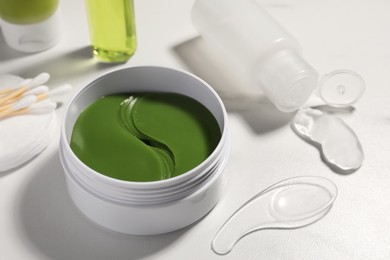 Photo of Jar of under eye patches with spoon and cosmetic products on white table, closeup