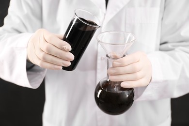 Photo of Woman pouring black crude oil from beaker into flask on dark background, closeup