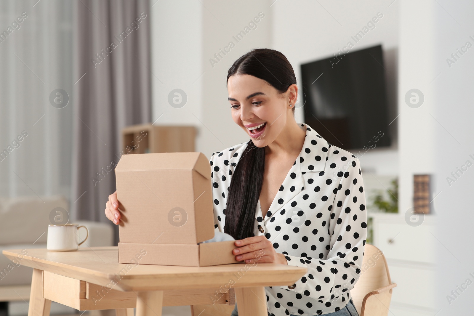 Photo of Happy young woman opening parcel at table indoors. Internet shopping