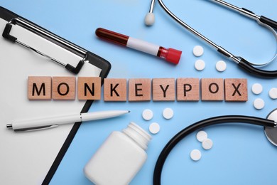 Photo of Word Monkeypox made of wooden cubes, test tube and pills on light blue background, flat lay