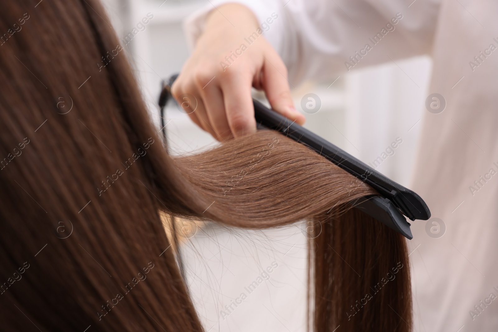 Photo of Hairdresser straightening woman's hair with flat iron indoors, closeup