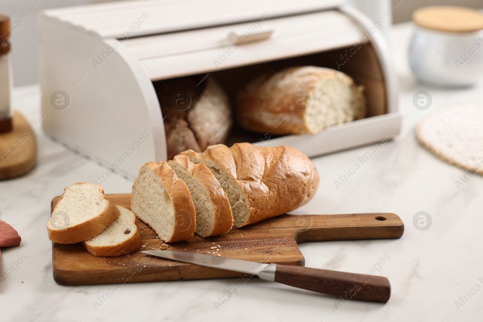 Photo of Wooden bread basket with freshly baked loaves and knife on white marble table in kitchen