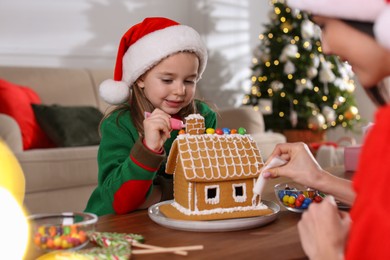 Photo of Mother and daughter decorating gingerbread house at table indoors