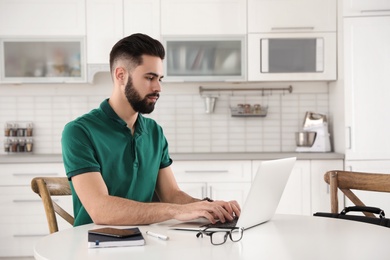 Photo of Handsome young man working with laptop at table in kitchen