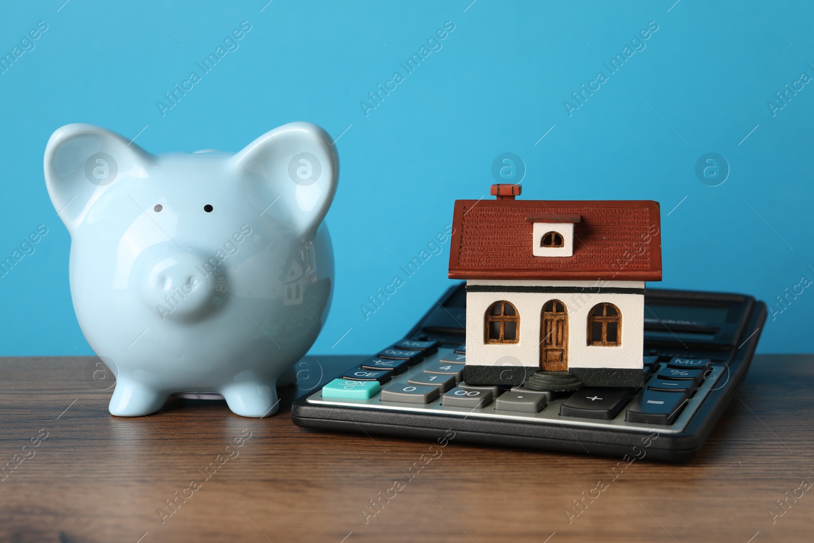 Photo of House model, piggy bank and calculator on wooden table against light blue background