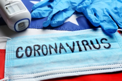 Photo of Word CORONAVIRUS written on protective mask, thermometer, gloves and American flag, closeup. Covid-19 pandemic in USA