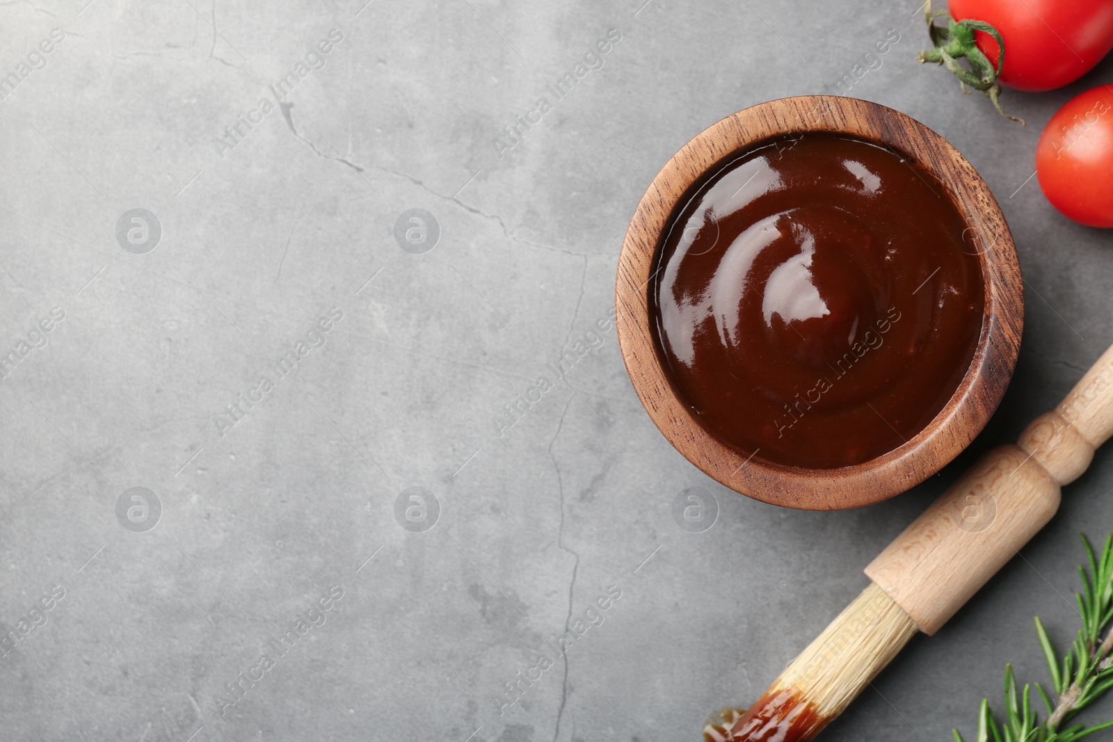 Photo of Tasty barbeque sauce in bowl, tomato, rosemary and brush on grey textured table, flat lay. Space for text