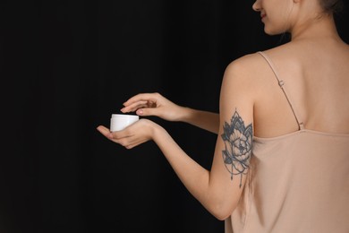 Photo of Woman with tattoo holding jar of cream against dark background, closeup