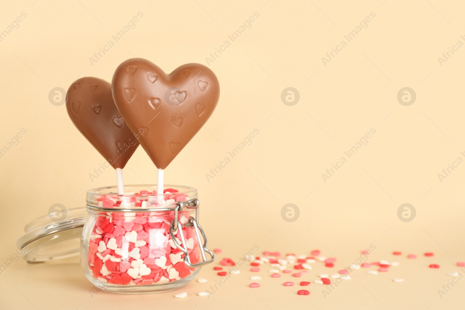 Photo of Chocolate heart shaped lollipops with sprinkles in glass jar on beige background. Space for text