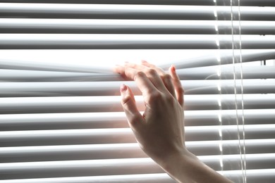 Photo of Woman separating slats of white blinds indoors, closeup
