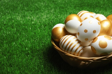 Photo of Wicker basket of traditional Easter eggs decorated with golden paint on green lawn, space for text