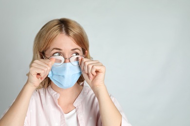 Woman wiping foggy glasses caused by wearing medical mask on light background. Space for text