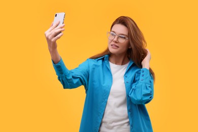 Beautiful woman in eyeglasses taking selfie on orange background