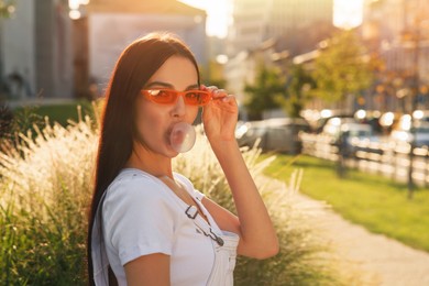 Beautiful woman in sunglasses blowing gum outdoors, space for text