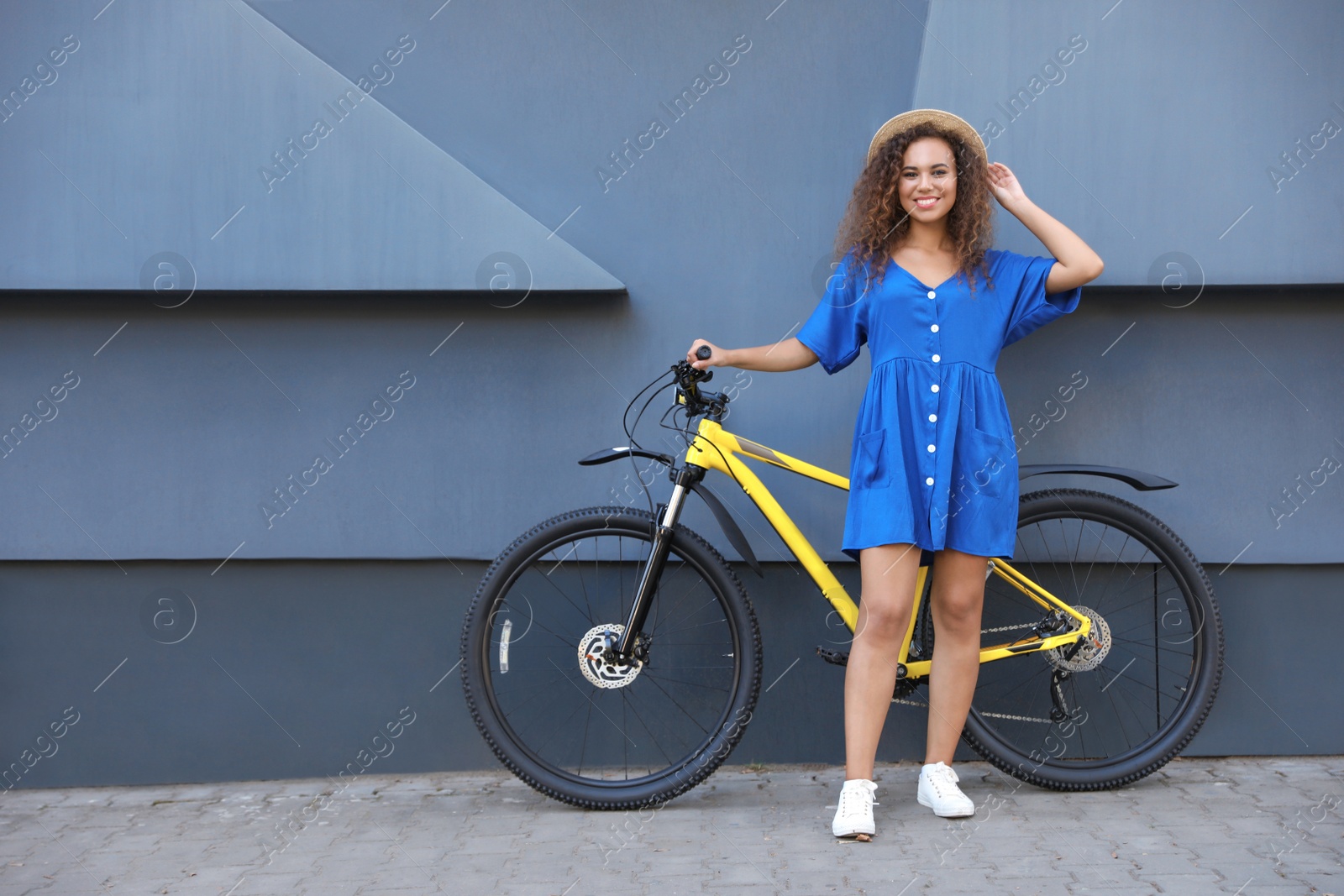 Photo of Beautiful young African-American woman with bicycle near grey wall on city street. Space for text