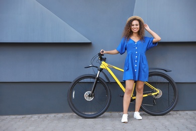 Beautiful young African-American woman with bicycle near grey wall on city street. Space for text