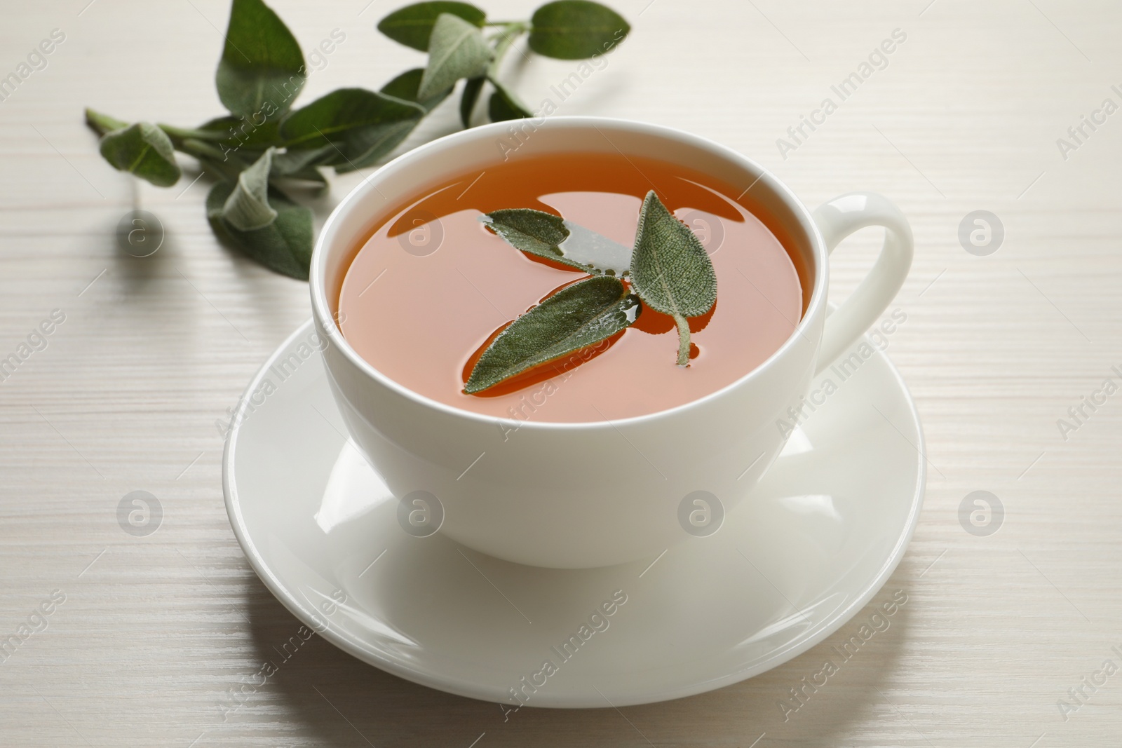 Photo of Cup of aromatic sage tea with fresh leaves on white wooden table