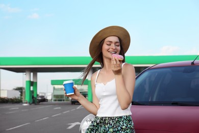 Photo of Beautiful young woman with coffee eating doughnut near car at gas station
