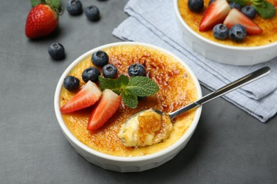 Photo of Delicious creme brulee with berries in bowl and spoon on grey table, closeup