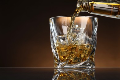 Pouring whiskey from bottle into glass with ice cubes at table against brown background, space for text