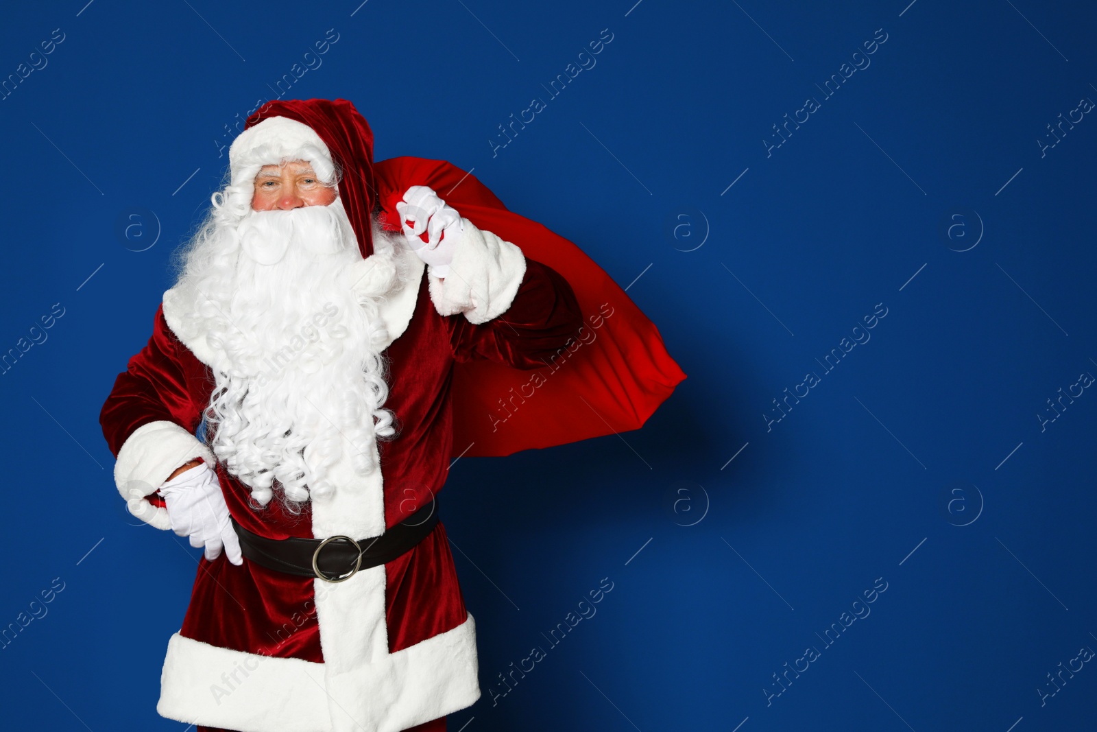 Photo of Happy authentic Santa Claus with bag full of gifts on blue background. Space for text