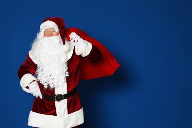 Photo of Happy authentic Santa Claus with bag full of gifts on blue background. Space for text