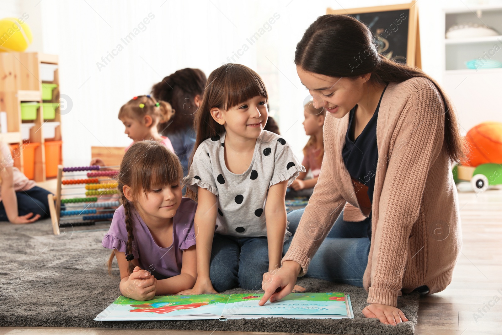 Photo of Kindergarten teacher reading book with cute girls while other children playing together indoors