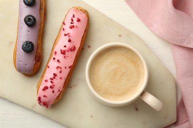 Photo of Cup of coffee and delicious eclairs covered with glaze on white wooden table, top view