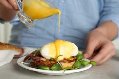 Photo of Woman pouring Hollandaise sauce onto egg Benedict at table, closeup