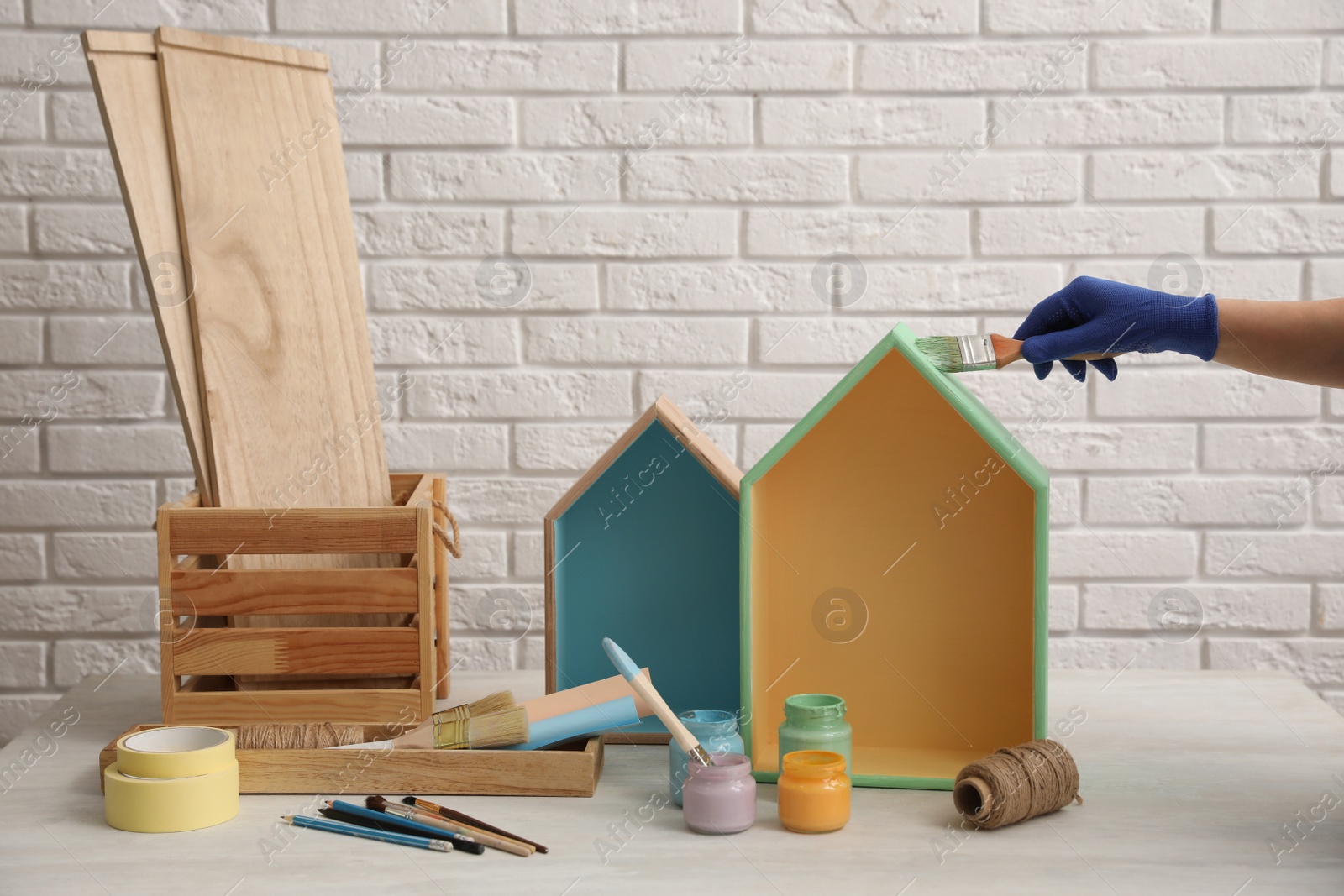 Photo of Decorator painting house shaped shelf with brush at white table, closeup. Interior element