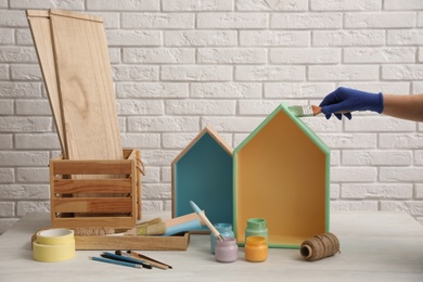 Photo of Decorator painting house shaped shelf with brush at white table, closeup. Interior element