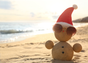 Photo of Snowman made of sand with Santa hat and sunglasses on beach near sea, space for text. Christmas vacation