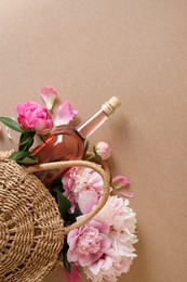 Photo of Wicker bag with bottle of rose wine and beautiful pink peonies on brown background, top view