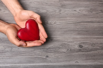 Young man holding red heart on grey wooden background, top view with space for text. Donation concept