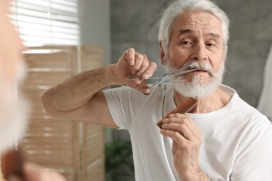 Photo of Senior man trimming mustache with scissors near mirror in bathroom