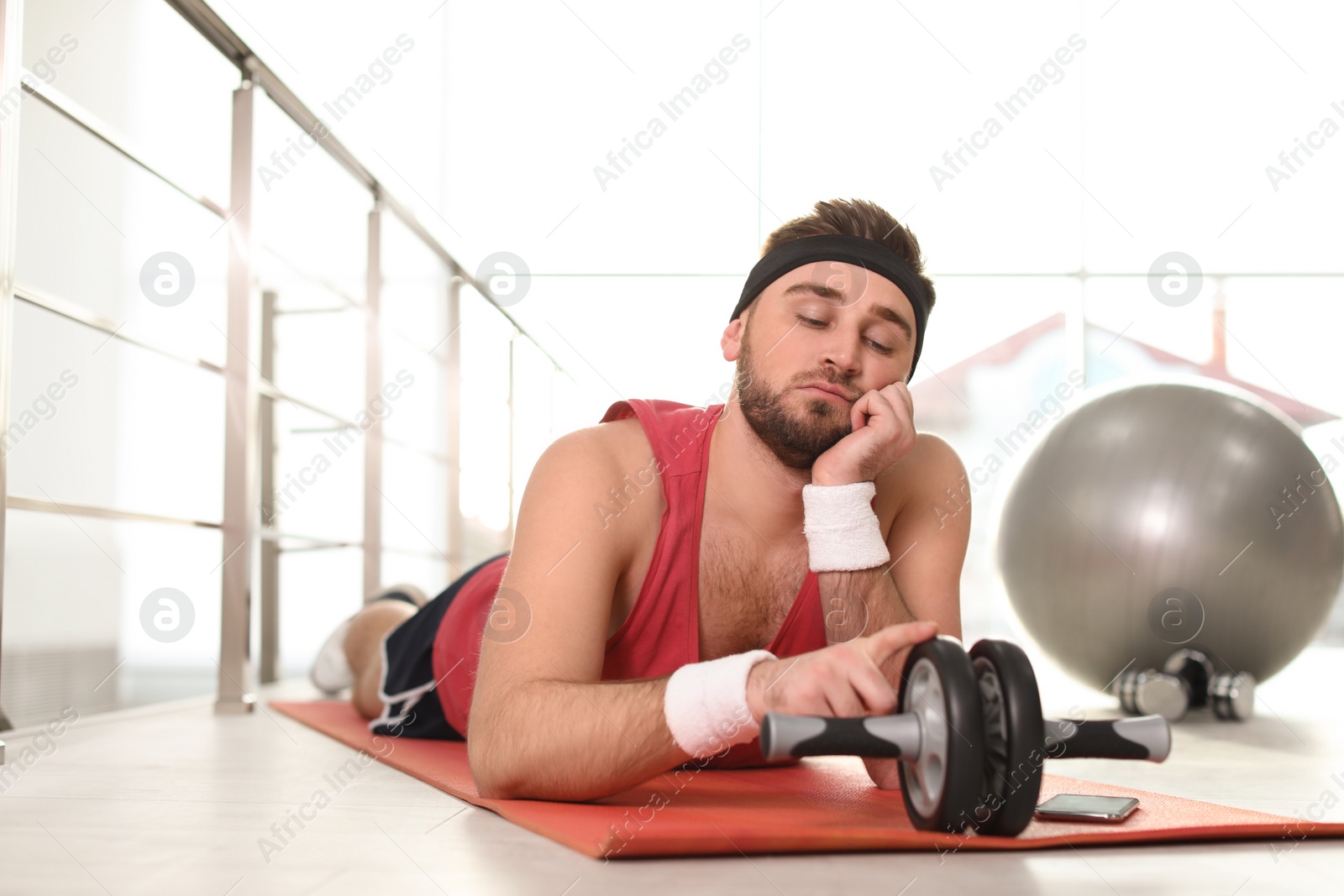 Photo of Lazy young man with abs roller lying on yoga mat indoors