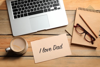 Card with phrase "I love Dad" near laptop and cup of coffee on wooden table, top view. Father's day celebration