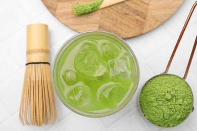 Photo of Glass of delicious iced green matcha tea, powder, bamboo whisk and spoon on white tiled table, flat lay