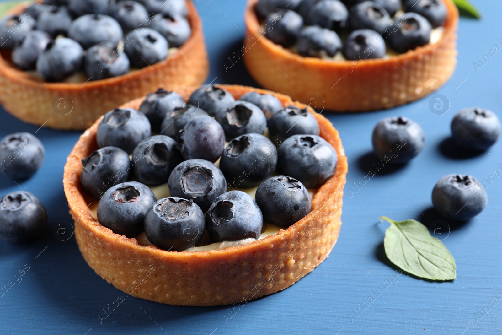 Photo of Tartlets with fresh blueberries and mint on blue table, closeup. Delicious dessert