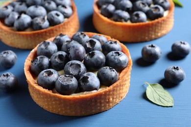 Photo of Tartlets with fresh blueberries and mint on blue table, closeup. Delicious dessert