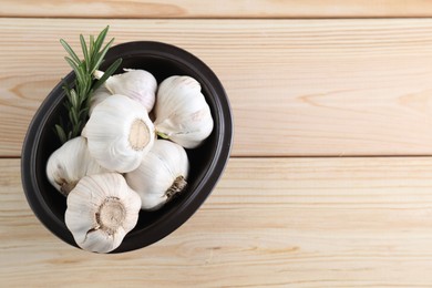 Photo of Fresh garlic bulbs in bowl and rosemary on wooden table, top view. Space for text